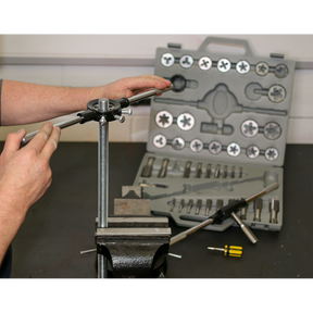 A person is using a threading tool on a metal rod, held in a vise. A Sealey Tap & Die Set 45pc Split Dies Metric - AK303, featuring various Metric sizes and Tungsten steel taps and dies, is open on the table in the background.