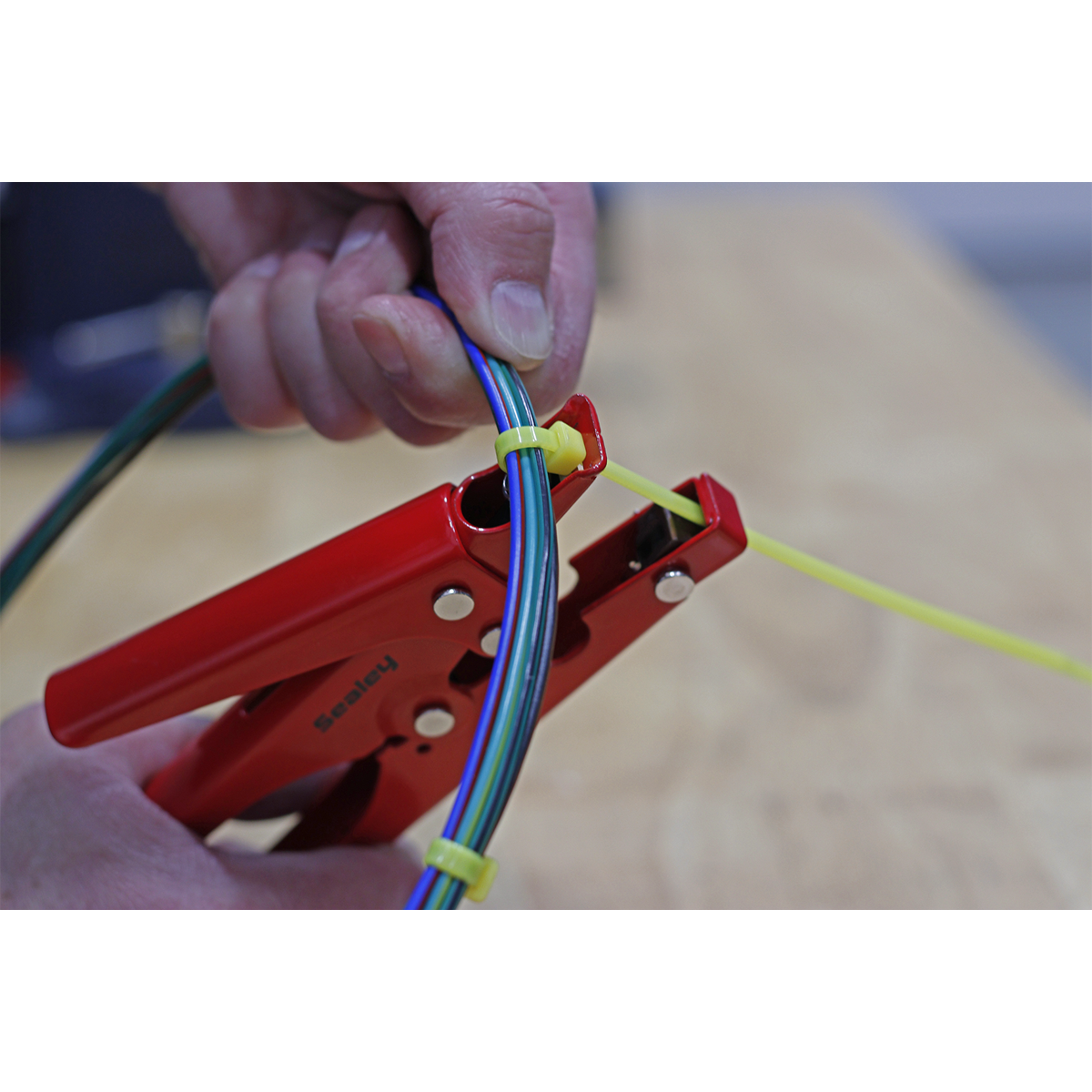 Person using the Sealey Cable Tie Fastening Tool - AK3254 with a spring-loaded handle to tighten a yellow cable tie around several multi-colored wires on a wooden surface.