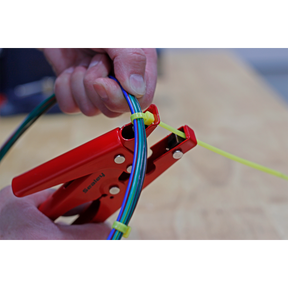 Person using the Sealey Cable Tie Fastening Tool - AK3254 with a spring-loaded handle to tighten a yellow cable tie around several multi-colored wires on a wooden surface.