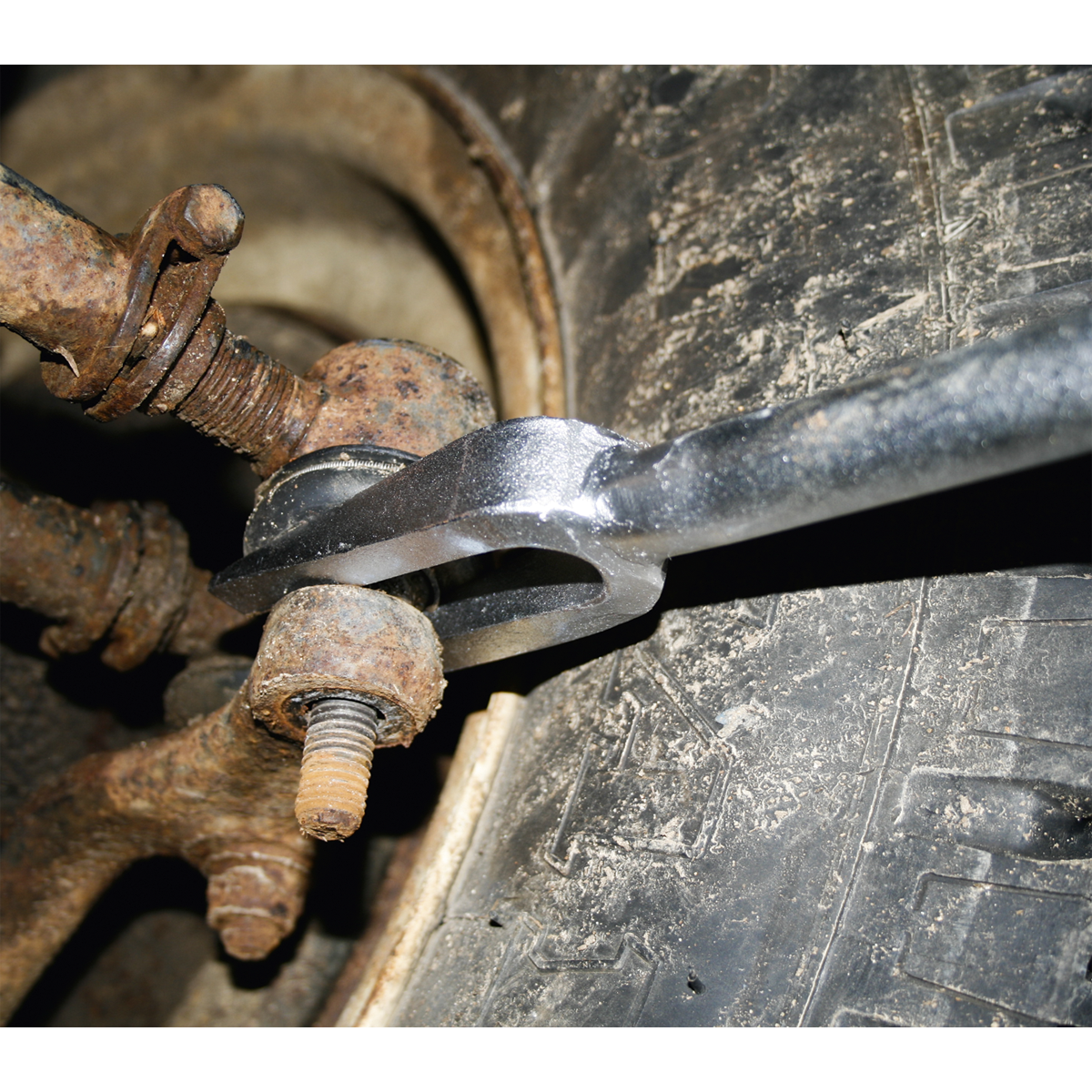 The Sealey Ball Joint Splitter Long Reach 400mm - AK3821 is being used to tighten a bolt on a rusty vehicle part near a tire with limited access.