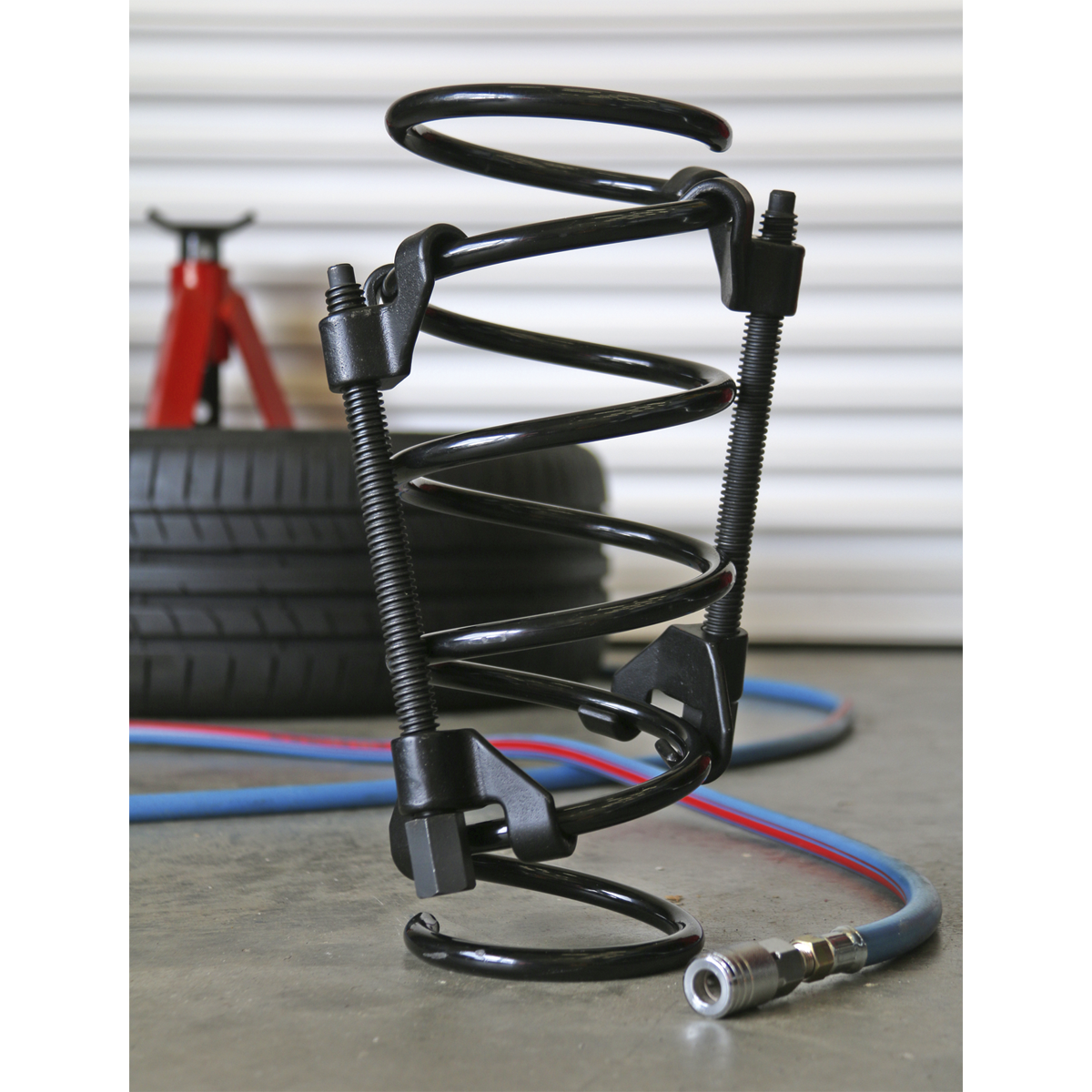 A Sealey Coil Spring Compressor 2pc - AK3841 rests on the floor beside a red jack, tires, and colored cables. The setup features hardened lead screws for durability.