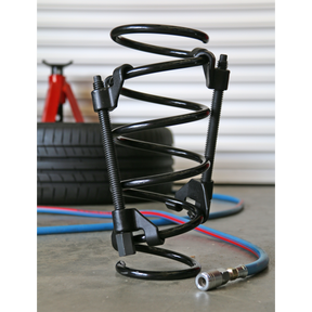A Sealey Coil Spring Compressor 2pc - AK3841 rests on the floor beside a red jack, tires, and colored cables. The setup features hardened lead screws for durability.