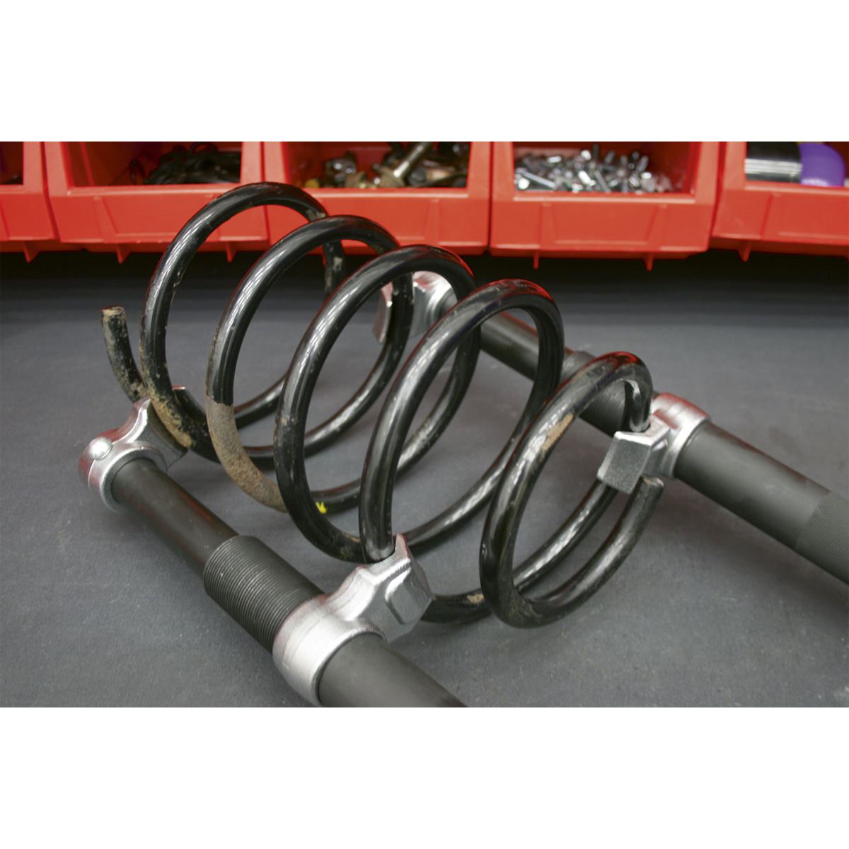 A close-up of the Sealey Coil Spring Compressor Set 2pc Heavy-Duty 2500kg/Pair - AK3845 on a workshop surface, with red storage bins containing various tools and parts in the background.