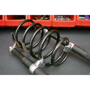 A close-up of the Sealey Coil Spring Compressor Set 2pc Heavy-Duty 2500kg/Pair - AK3845 on a workshop surface, with red storage bins containing various tools and parts in the background.