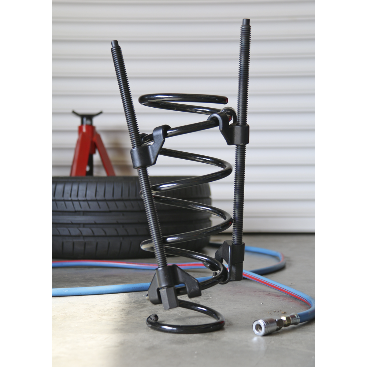 A close-up of a car suspension coil spring compressed by the robust Sealey Coil Spring Compressor 2pc Long Reach - AK3846, with long reach jaws ensuring precision. In the background, tires, a red jack stand, and an air hose complete the scene.