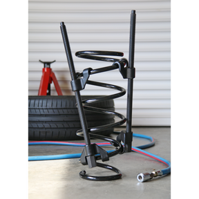 A close-up of a car suspension coil spring compressed by the robust Sealey Coil Spring Compressor 2pc Long Reach - AK3846, with long reach jaws ensuring precision. In the background, tires, a red jack stand, and an air hose complete the scene.
