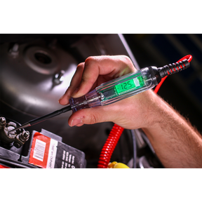 A person uses a Sealey Integrated Test Light/Voltmeter 3-48V - AK4012 to measure the voltage of a car battery, displaying a reading of 12.5 volts.