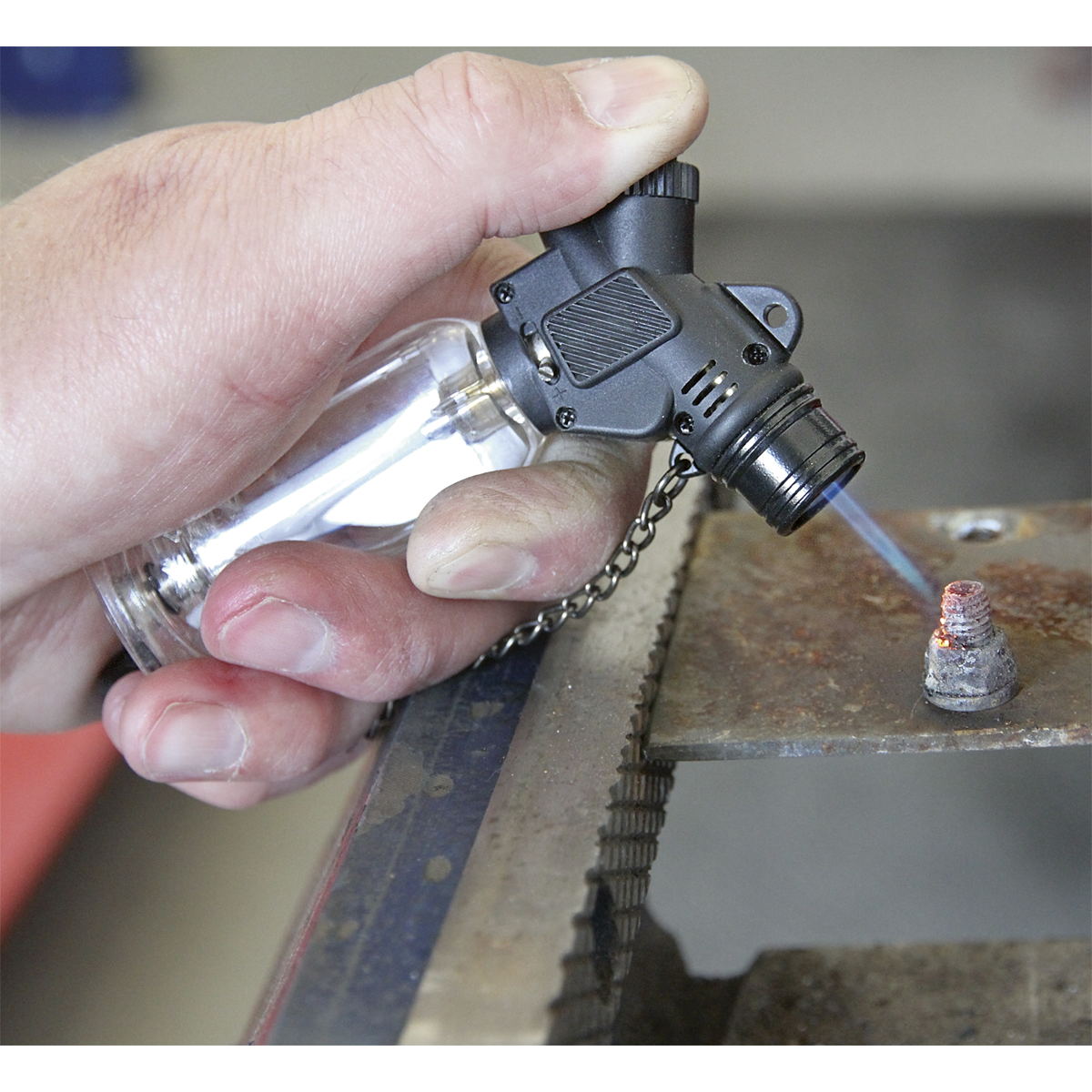 A hand holds the Sealey Butane Micro Heating Torch - AK4042 with adjustable flame control, directing a blue flame at a metal object on a workbench, showcasing the precision of professional hand tools.