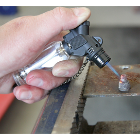 A hand holds the Sealey Butane Micro Heating Torch - AK4042 with adjustable flame control, directing a blue flame at a metal object on a workbench, showcasing the precision of professional hand tools.