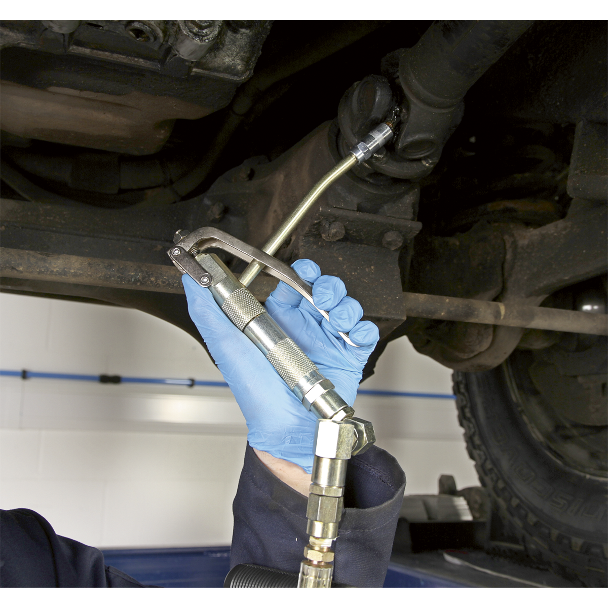 A person wearing blue gloves uses the Sealey Grease Pump Air Operated 50kg - AK453X connected to a grease hose to lubricate a component underneath a vehicle.