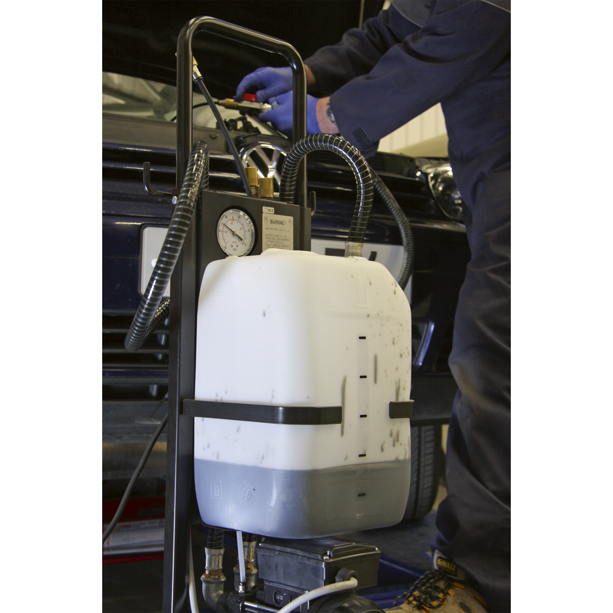 A technician wearing gloves efficiently services a vehicle using the Sealey Oil Extractor Mobile 230V - AK466D. In the foreground, a white container equipped with tubes and gauges ensures spill-free oil removal.