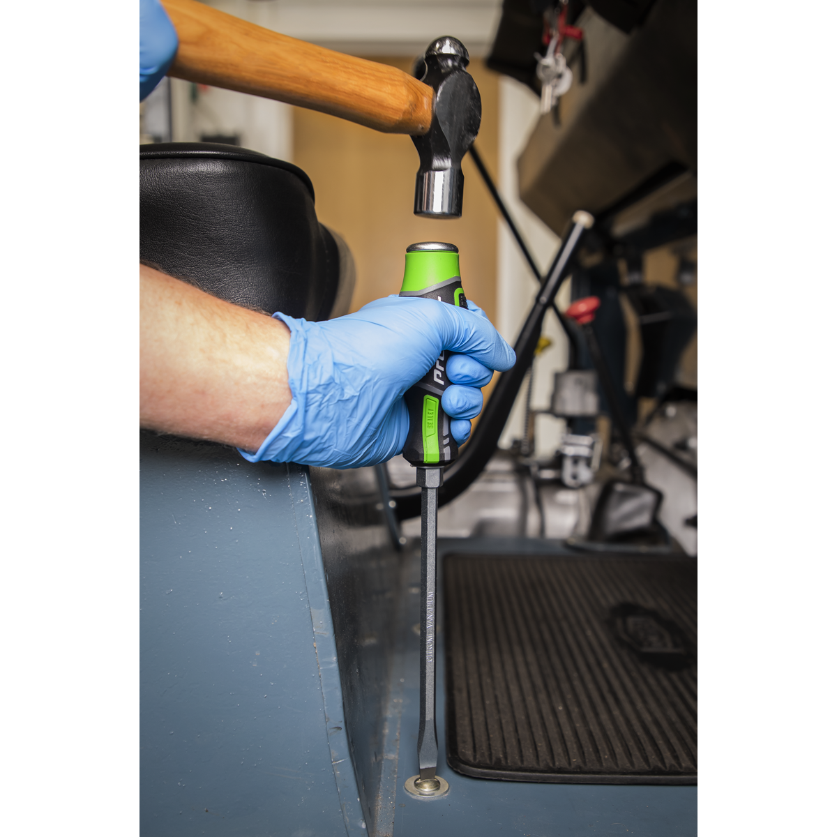 A person wearing blue gloves grips a soft-handled Premier Hand Tool from the Sealey Hammer-Thru Screwdriver Set Hi-Vis Green 6pc - AK4940HV against a metal surface while striking it with a hammer.