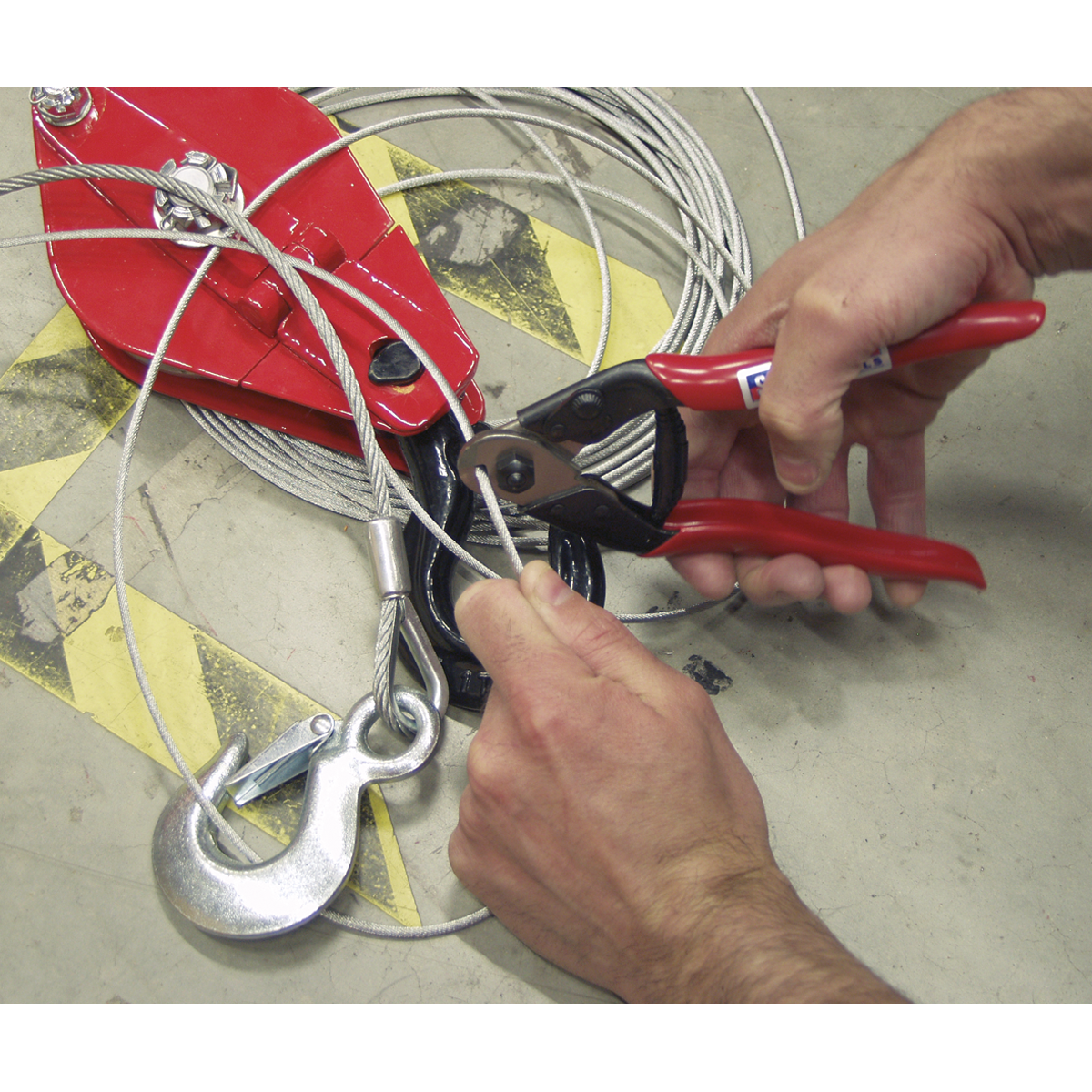 Close-up of hands using a Sealey Wire Rope/Spring Cutter 190mm - AK503 with high carbon steel blades to trim a wire rope near a large metal hook and pulley system.