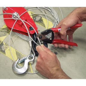 Close-up of hands using a Sealey Wire Rope/Spring Cutter 190mm - AK503 with high carbon steel blades to trim a wire rope near a large metal hook and pulley system.