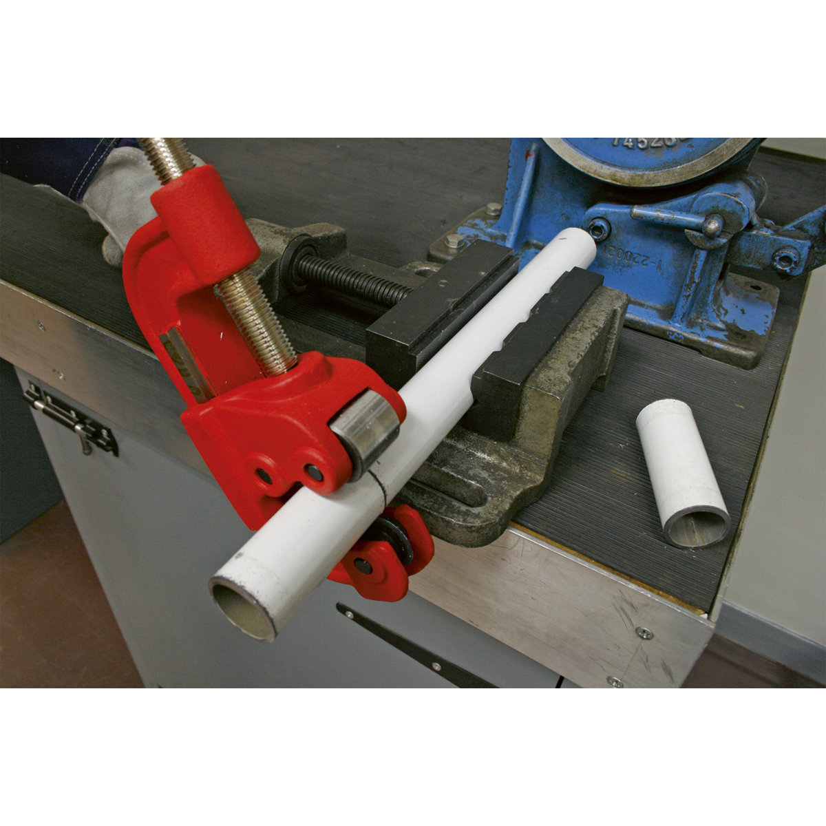 A white pipe is being cut with the Sealey Pipe Cutter Ø10-50mm Capacity - AK5062, secured in a bench vise on a workbench. The hand tool's hardened steel blade ensures a clean cut. Nearby, a piece of the cut pipe lies neatly on the surface.