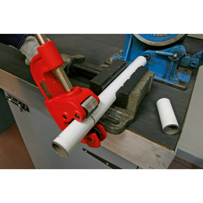 A white pipe is being cut with the Sealey Pipe Cutter Ø10-50mm Capacity - AK5062, secured in a bench vise on a workbench. The hand tool's hardened steel blade ensures a clean cut. Nearby, a piece of the cut pipe lies neatly on the surface.