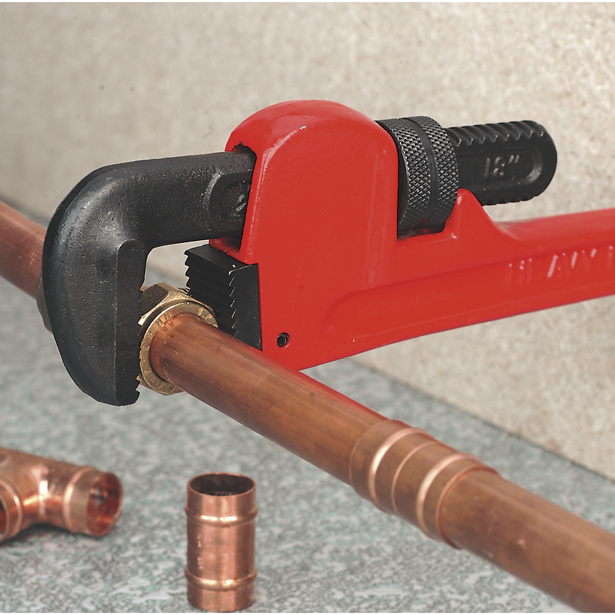 A Sealey Pipe Wrench European Pattern 250mm Cast Steel - AK5102, featuring high-strength, drop-forged carbon steel jaws, is being used to tighten a brass fitting on a copper pipe. Other pieces of copper piping are scattered on a gray surface. Trust in the reliability of Sealey Hand Tools, which come with a lifetime guarantee.