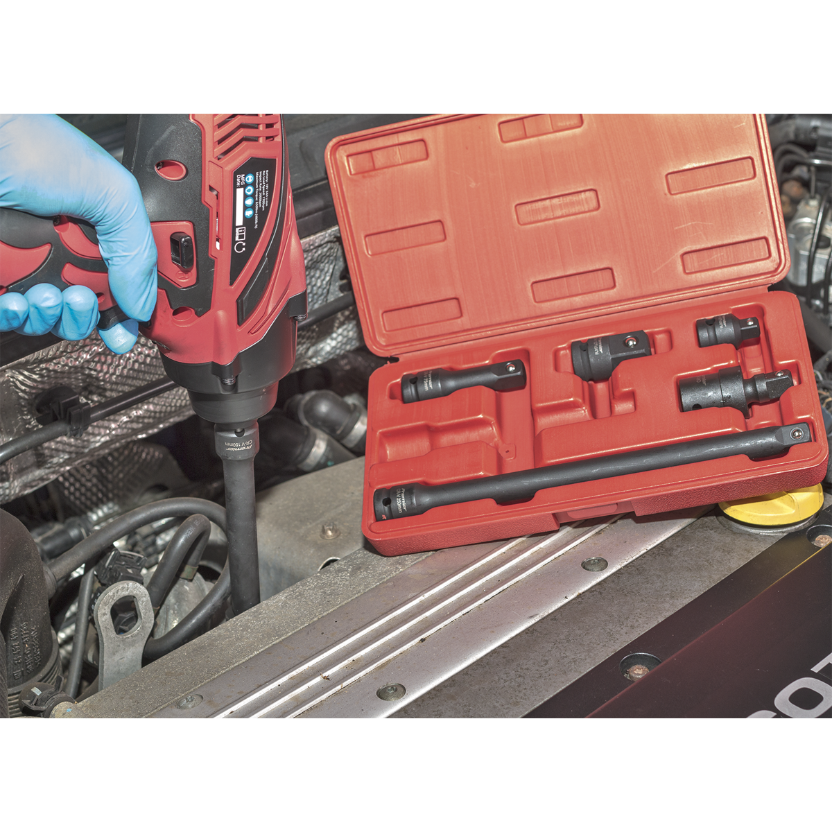 A person wearing a blue glove uses a power drill on a car engine. Next to the engine, an open red case containing the Sealey Premier 6pc Impact Adaptor & Extension Bar Set 1/2"Sq Drive (model AK5514) and various attachments is visible.