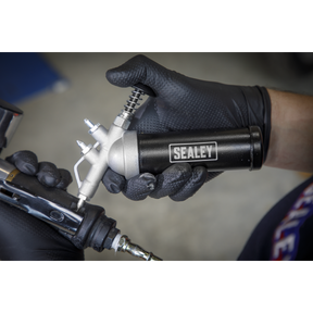 A person wearing black gloves holds and uses the Sealey Mini Grease Gun Push Type - AK55, demonstrating precise placement for optimal efficiency.