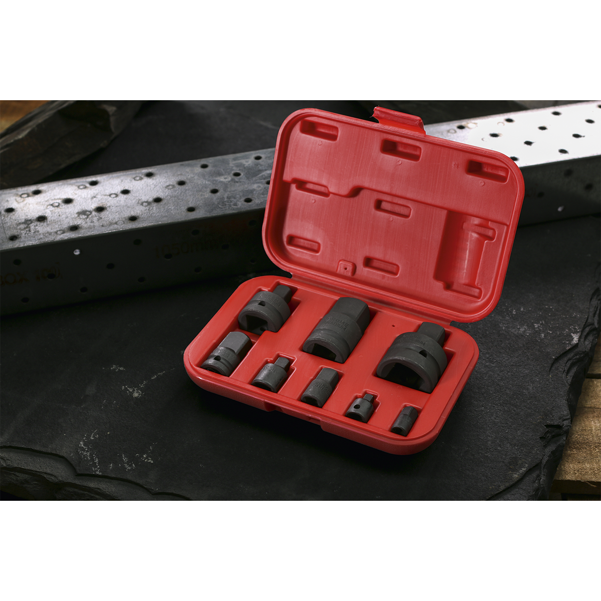 A red plastic case from Sealey, specifically the Impact Socket Adaptor Set 8pc - AK5900B, is open and displayed on a dark surface. The set includes various black metal hole saws of different sizes and impact adaptors made from durable Chrome Vanadium steel to ensure longevity and performance.