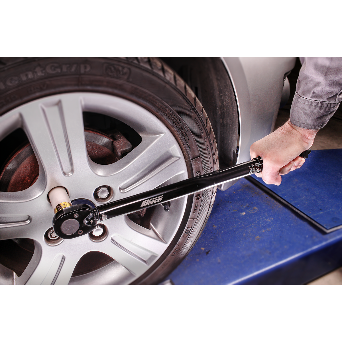 A person is using a Sealey Micrometer Torque Wrench 1/2"Sq Drive Calibrated Black Series - AK624B to tighten the lug nuts on a car wheel on a lift in an automotive workshop, ensuring precise calibration tolerance with professional hand tools.