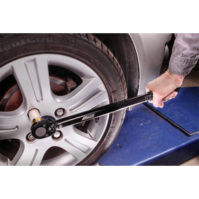 A person is using a Sealey Micrometer Torque Wrench 1/2"Sq Drive Calibrated Black Series - AK624B to tighten the lug nuts on a car wheel on a lift in an automotive workshop, ensuring precise calibration tolerance with professional hand tools.