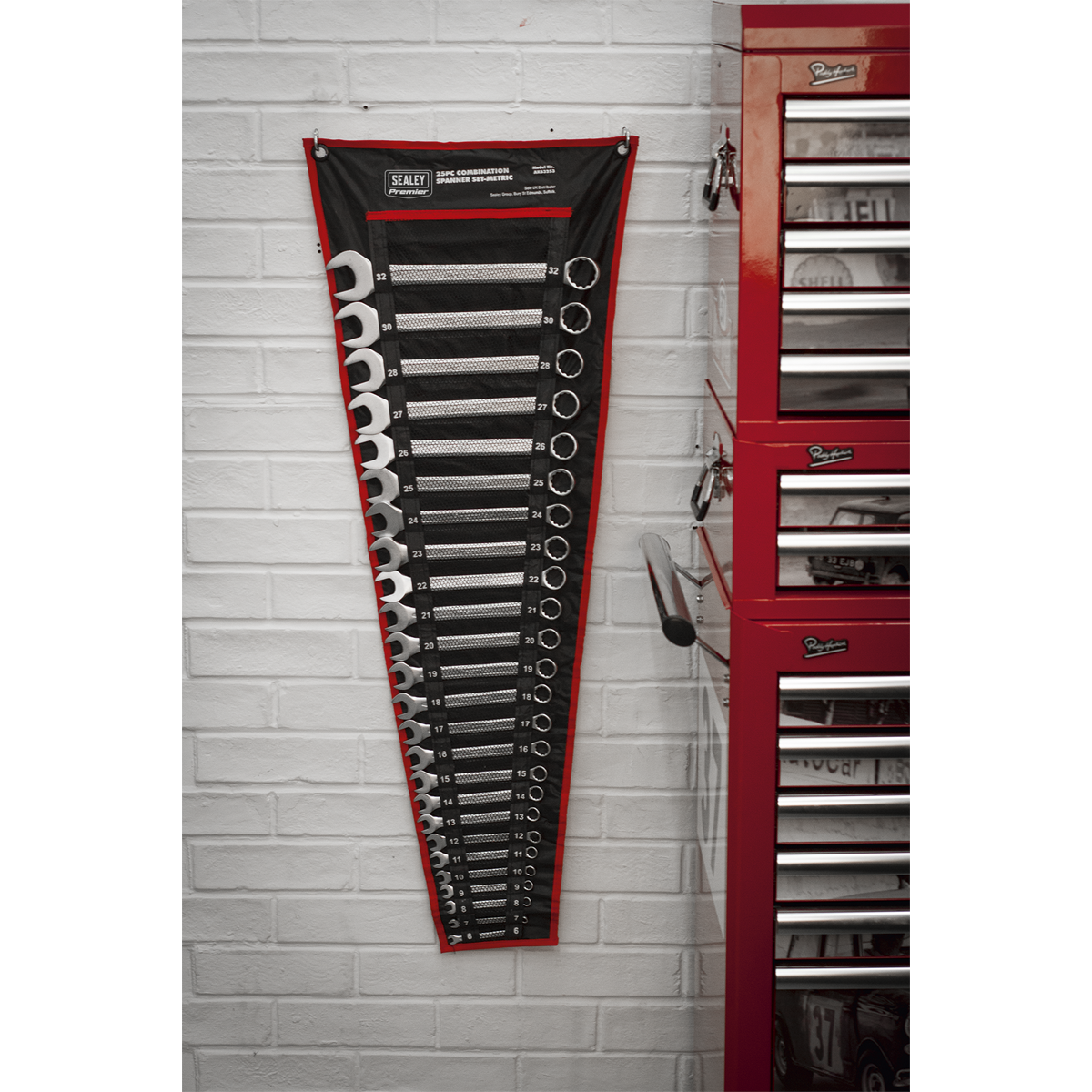 A wall-mounted Sealey Combination Spanner Set 25pc Metric - AK63253, featuring 12-point WallDrive® ring combination spanners, hangs next to a red tool chest in a workshop with white brick walls.