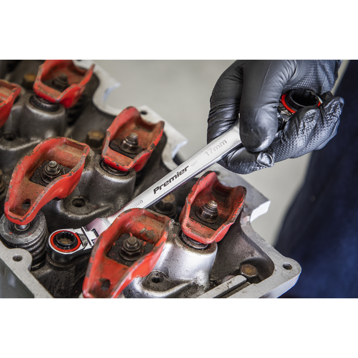 A gloved hand using a Sealey Ratchet Ring Spanner 4-in-1 Flexi-Head Reversible Metric Platinum Series - AK63947 made from Chrome Vanadium steel on a mechanical component with red and rusted parts.