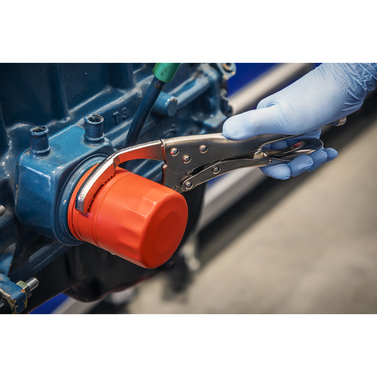 A gloved hand uses Sealey's Ø45-130mm Oil Filter Locking Pliers - Angled (AK6423) to tighten a red oil filter on a blue engine component.