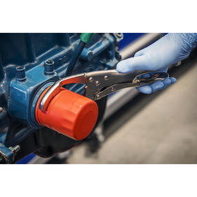 A gloved hand uses Sealey's Ø45-130mm Oil Filter Locking Pliers - Angled (AK6423) to tighten a red oil filter on a blue engine component.