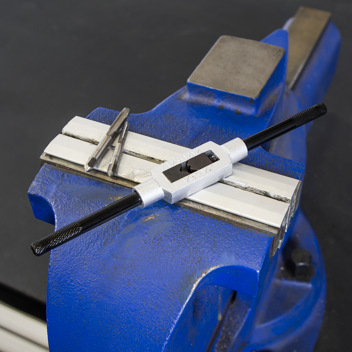 A blue bench vise holds a Sealey Tap Wrench M3-M12 - AK727 with sliding jaws and two black handles, designed for threading holes. Next to the wrench, two taper taps labeled M3 and M12 lie nearby.