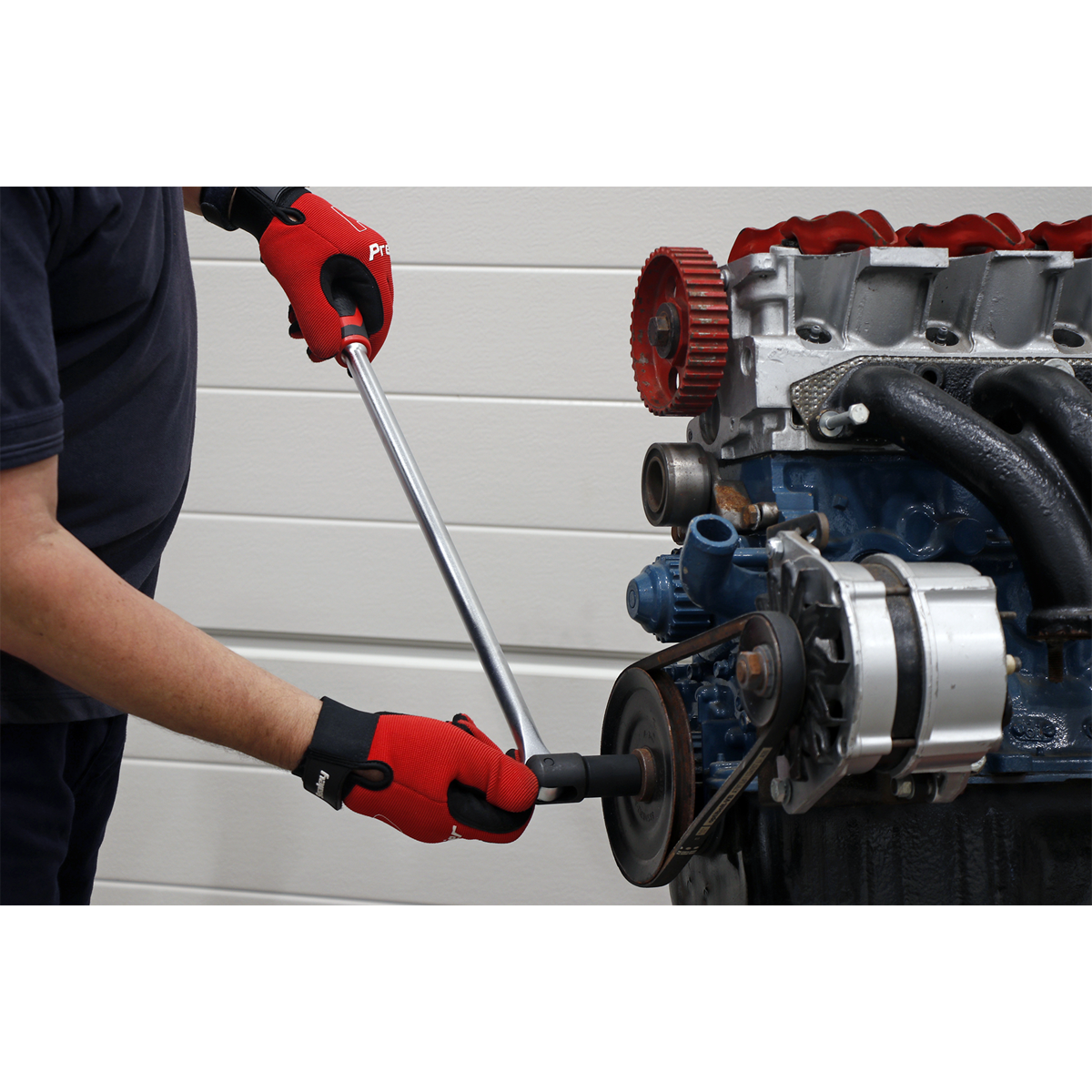 A person wearing red gloves with an anti-slip comfort grip uses a Sealey Breaker Bar 600mm 1/2" Sq Drive - AK7304 on a car engine belt system against a white wall background.