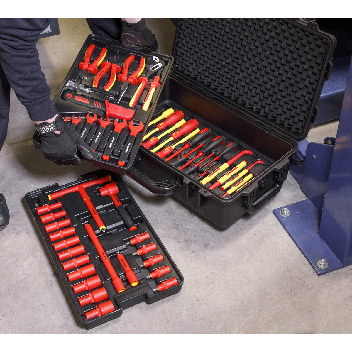 A person is meticulously organizing the Sealey 1000V Insulated Tool Kit 3/8"Sq Drive 50pc - AK7938, which includes pliers, screwdrivers, and sockets in red and yellow, in black plastic cases on a concrete floor for professional use.