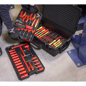 A person is meticulously organizing the Sealey 1000V Insulated Tool Kit 3/8"Sq Drive 50pc - AK7938, which includes pliers, screwdrivers, and sockets in red and yellow, in black plastic cases on a concrete floor for professional use.