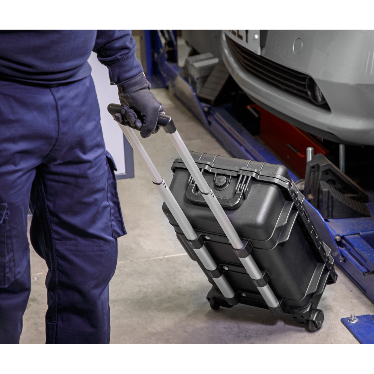 A person in blue overalls pulls a black hard shell case with a telescopic handle on wheels, potentially containing the Sealey 1000V Insulated Tool Kit 1/2"Sq Drive 49pc - AK7939, in an industrial or workshop setting.