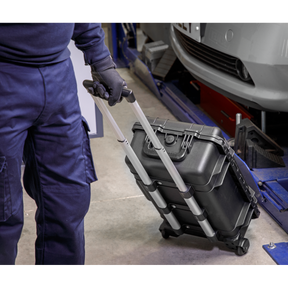 A person in blue overalls pulls a black hard shell case with a telescopic handle on wheels, potentially containing the Sealey 1000V Insulated Tool Kit 1/2"Sq Drive 49pc - AK7939, in an industrial or workshop setting.