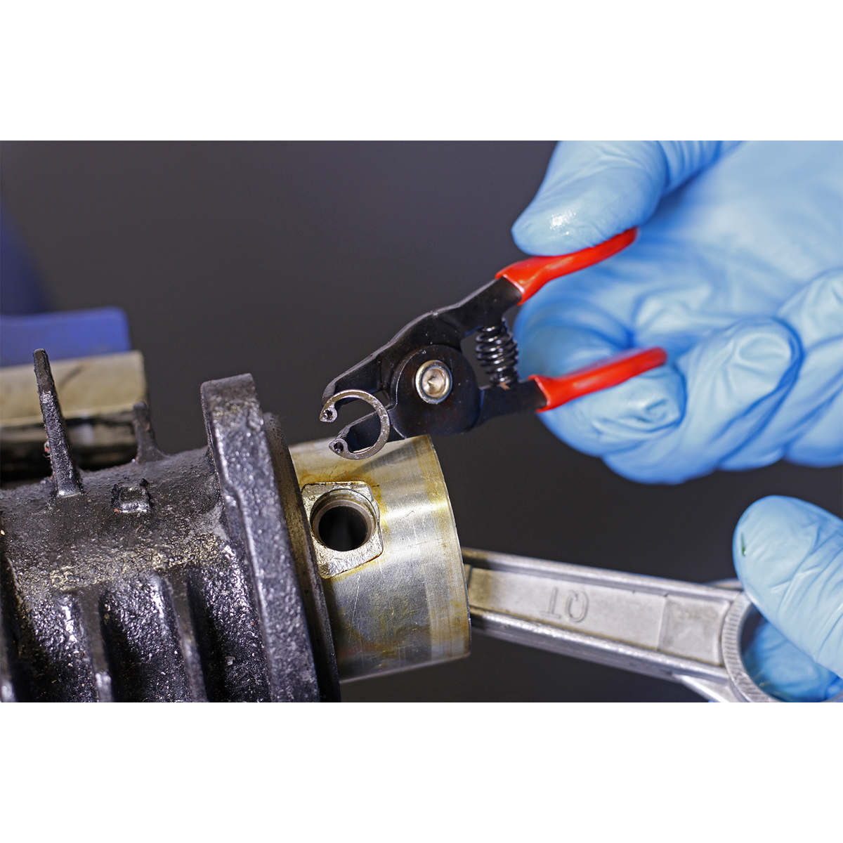Close-up of gloved hands using a pair from the Sealey Mini Circlip Pliers Set 4pc - AK8449 to remove a circlip from a mechanical component, held in place with a wrench. The precision and durability of these professional hand tools ensure efficient work.