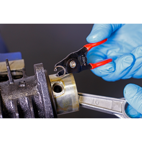 Close-up of gloved hands using a pair from the Sealey Mini Circlip Pliers Set 4pc - AK8449 to remove a circlip from a mechanical component, held in place with a wrench. The precision and durability of these professional hand tools ensure efficient work.