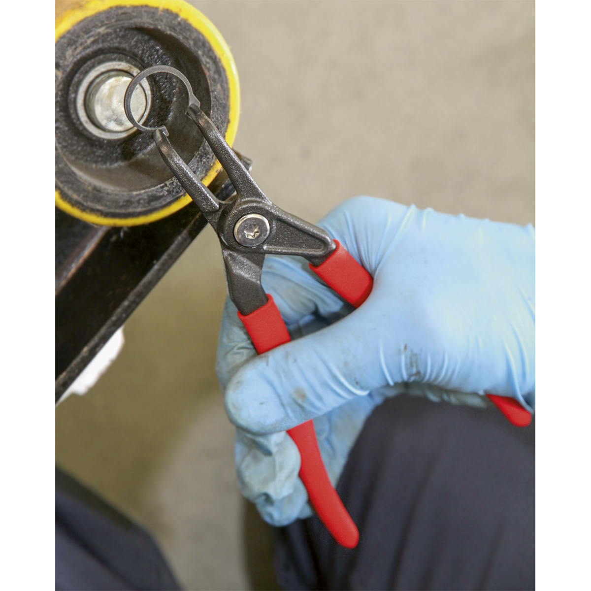 A person wearing blue gloves uses a pair of Sealey Circlip Pliers External Bent Nose 170mm, featuring red handles and crafted from high grade carbon steel, to adjust a round black component.