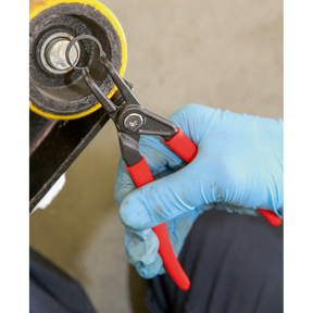 A person wearing blue gloves uses a pair of Sealey Circlip Pliers External Bent Nose 170mm, featuring red handles and crafted from high grade carbon steel, to adjust a round black component.