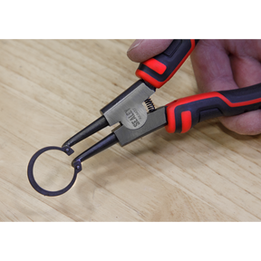 A hand holding the Sealey Circlip Pliers from the 180mm 4-piece set (AK8457) grips a circular metal ring on a wooden surface, showcasing the quality of Premier Hand Tools.