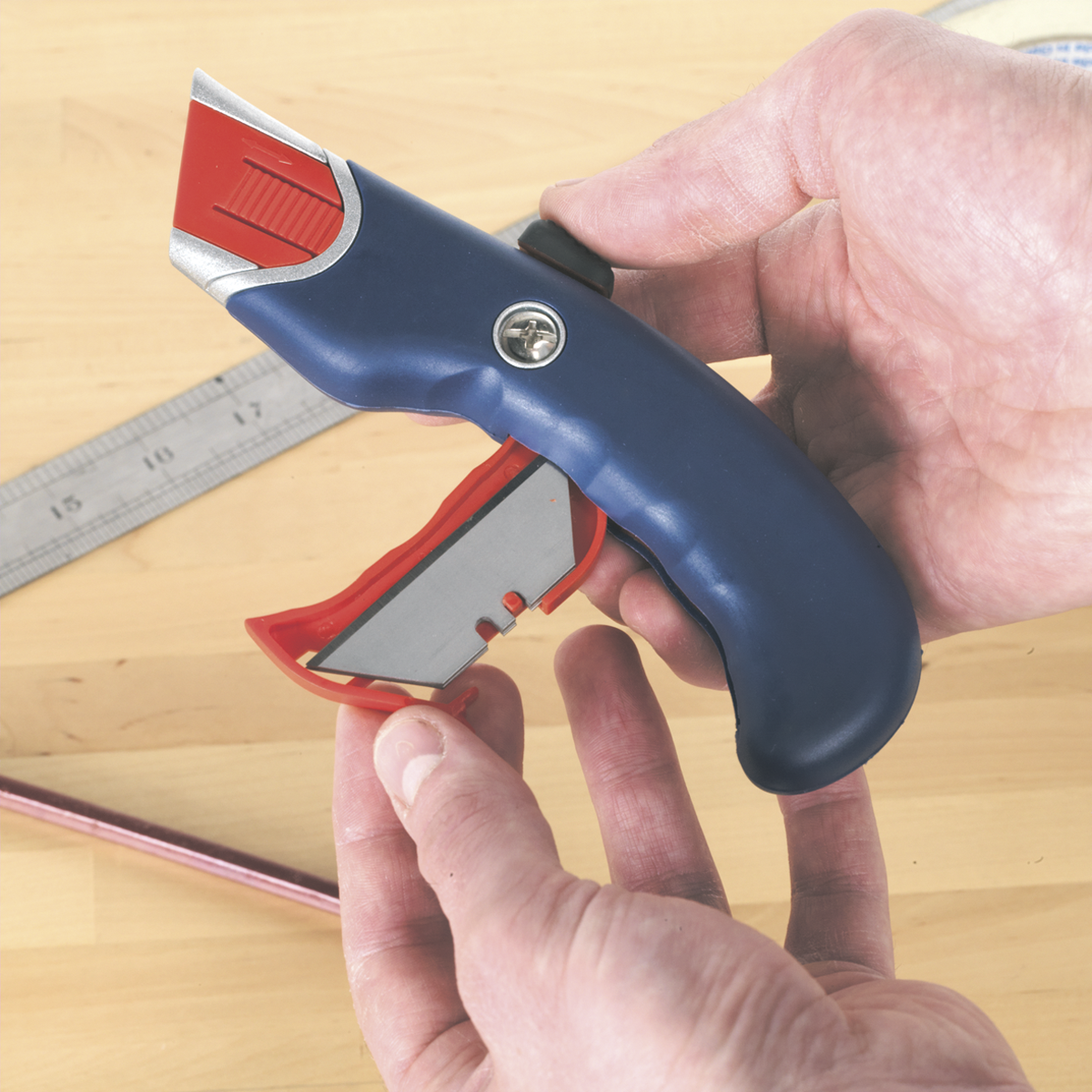 Hands holding a blue Sealey Safety Knife Auto-Retracting - AK8631 with an ergonomically profiled zinc alloy body, the blade compartment open to reveal a spare blade. A ruler and pencil lie on the wooden surface below, showcasing one of Sealey's finest creations.