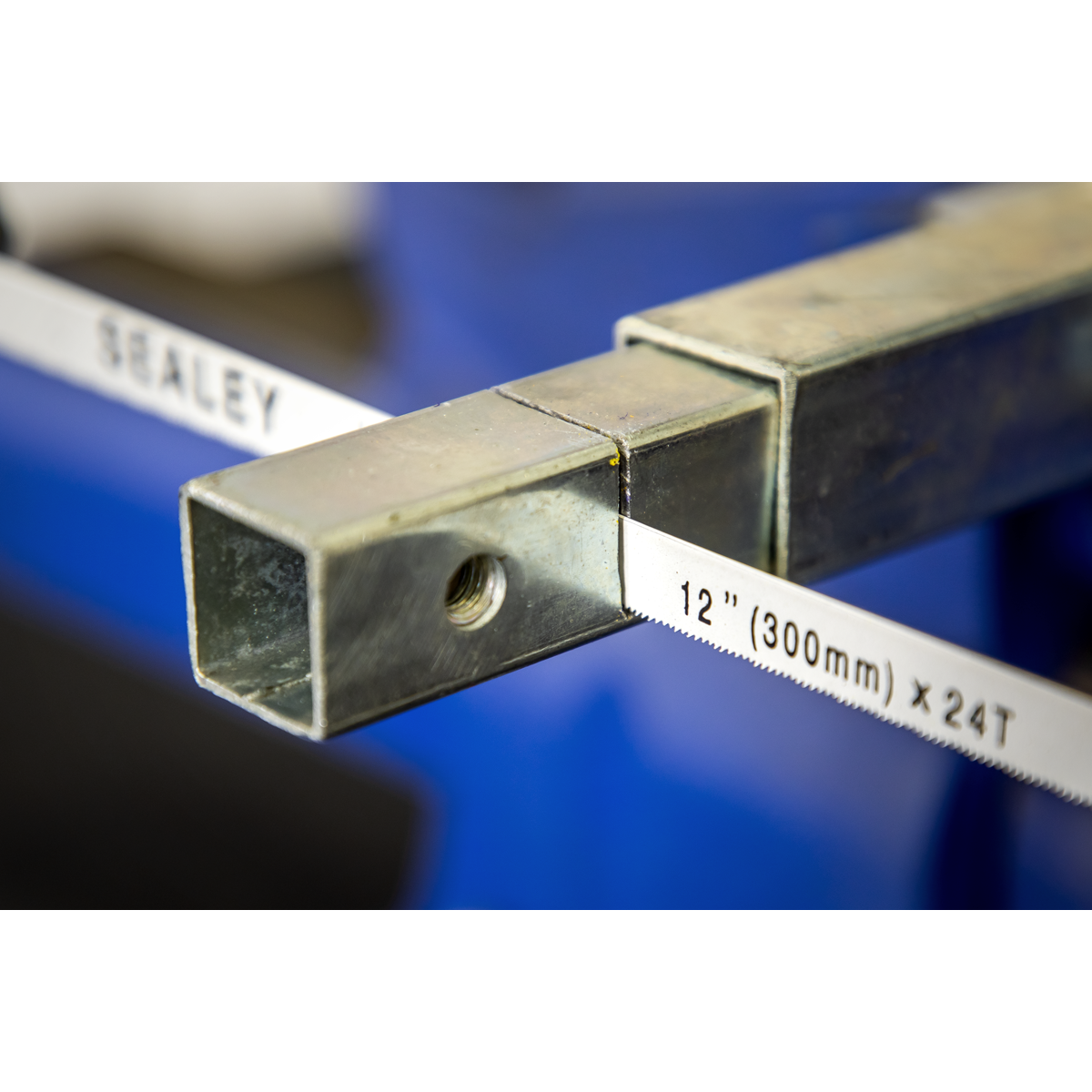 Close-up of a Sealey Hacksaw 300mm Professional - AK869 with a die-cast handle, cutting through a metal square tube clamped in a blue vise.