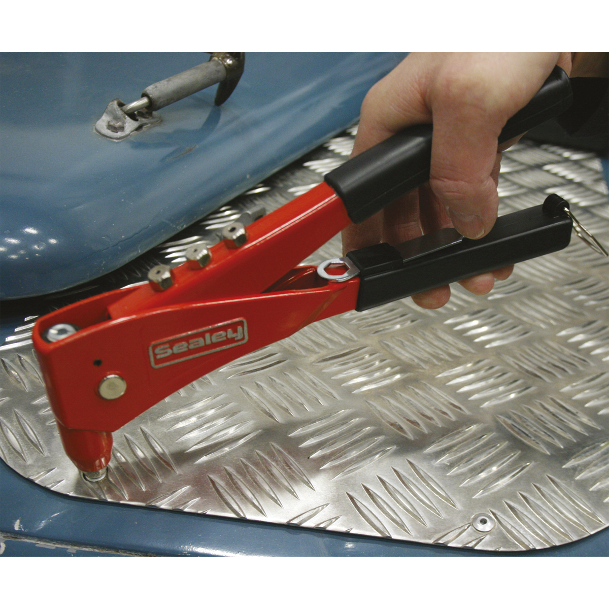 A hand using a red Sealey Hand Riveter Kit - AK99 with chrome steel jaws to apply an aluminum rivet to a blue metal surface with a diamond plate pattern.