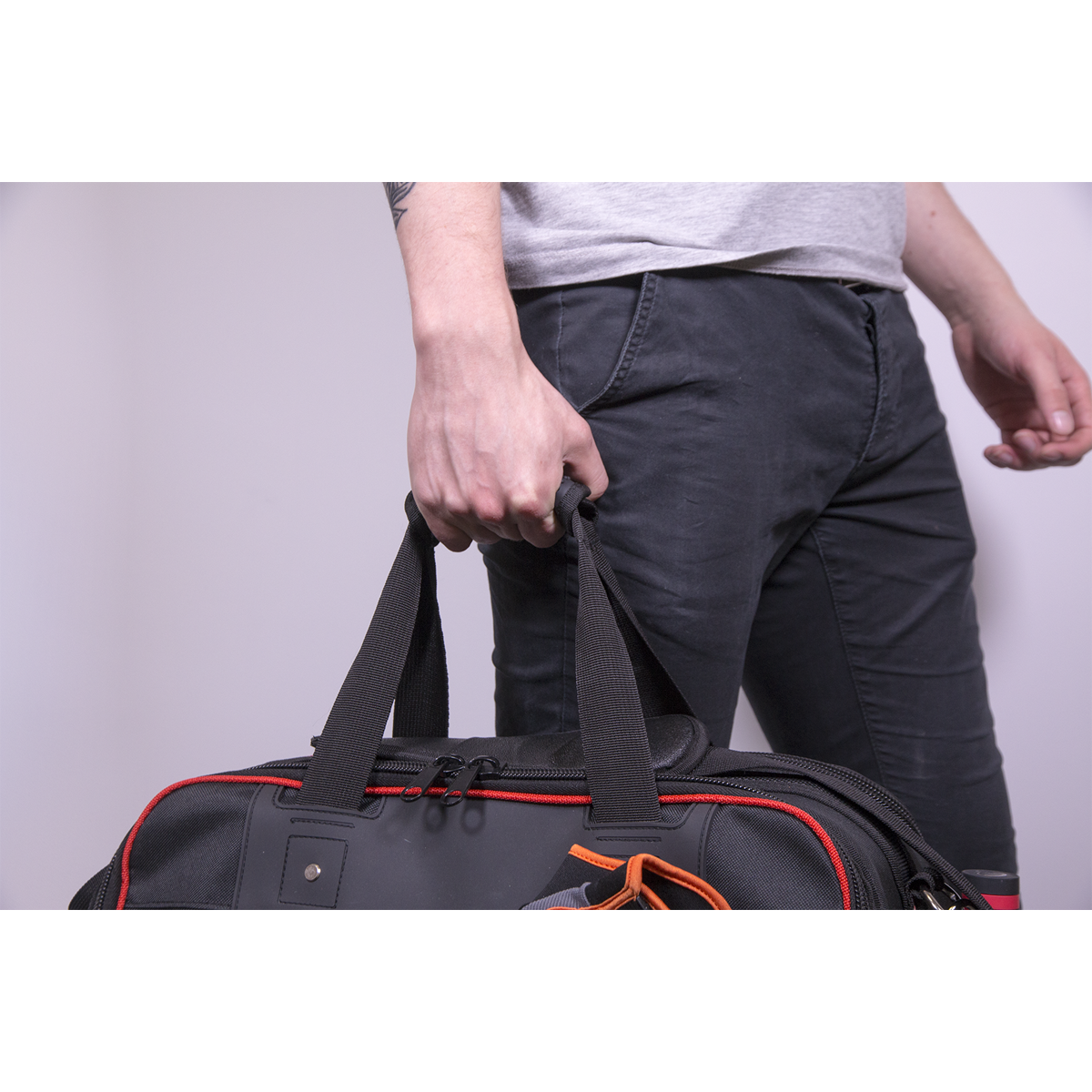 A person wearing dark pants and a light shirt holds the Sealey Tool Storage Bag with 24 Pockets 500mm Heavy-Duty - AP513, its black and red design standing out as they grasp its handles. Only the lower half of their body is visible.