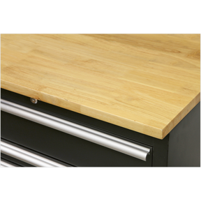 A close-up view of a Sealey Hardwood Worktop 1550mm - APMS07 with a smooth surface and the top of metal handles on black drawers beneath it, showcasing the details of Model No's APMS02 floor cabinets.