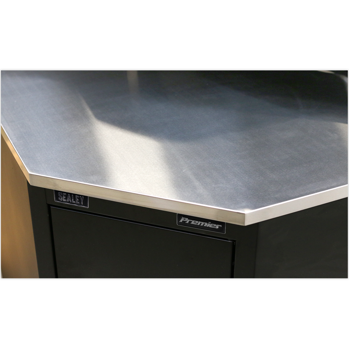 Close-up of a corner of a metallic workbench with "Sealey" label on a dark-colored cabinet, featuring a stainless steel worktop and Model No. APMS19 Stainless Steel Corner Worktop 930mm.