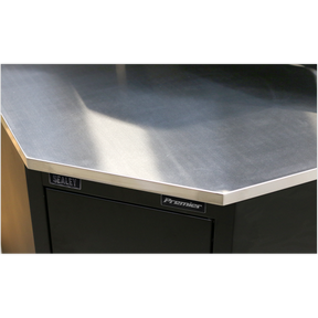Close-up of a corner of a metallic workbench with "Sealey" label on a dark-colored cabinet, featuring a stainless steel worktop and Model No. APMS19 Stainless Steel Corner Worktop 930mm.