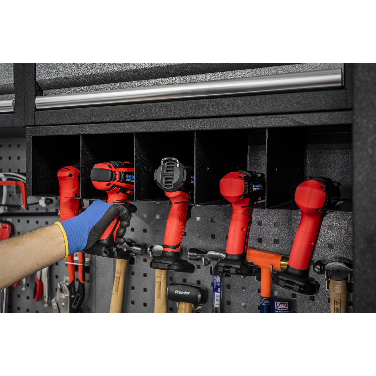 A hand reaches for red power tools stored in a black storage unit mounted on a pegboard, with other tools hanging below. Nearby, Sealey's APMS wall cabinets and the Modular Power Tool Rack 680mm - APMS69 offer additional power tool storage solutions.