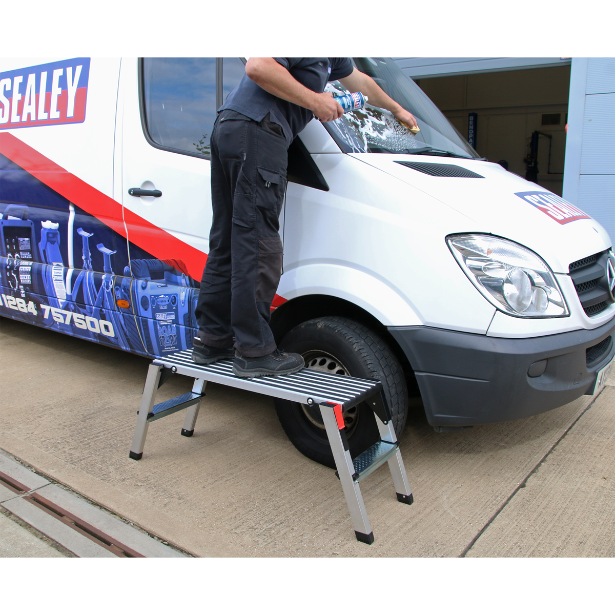 An individual stands on a Sealey Aluminium Folding Platform 2-Tread EN 131-4 (APS2) while cleaning the windshield of a white commercial van with a billboard-style design on its side.